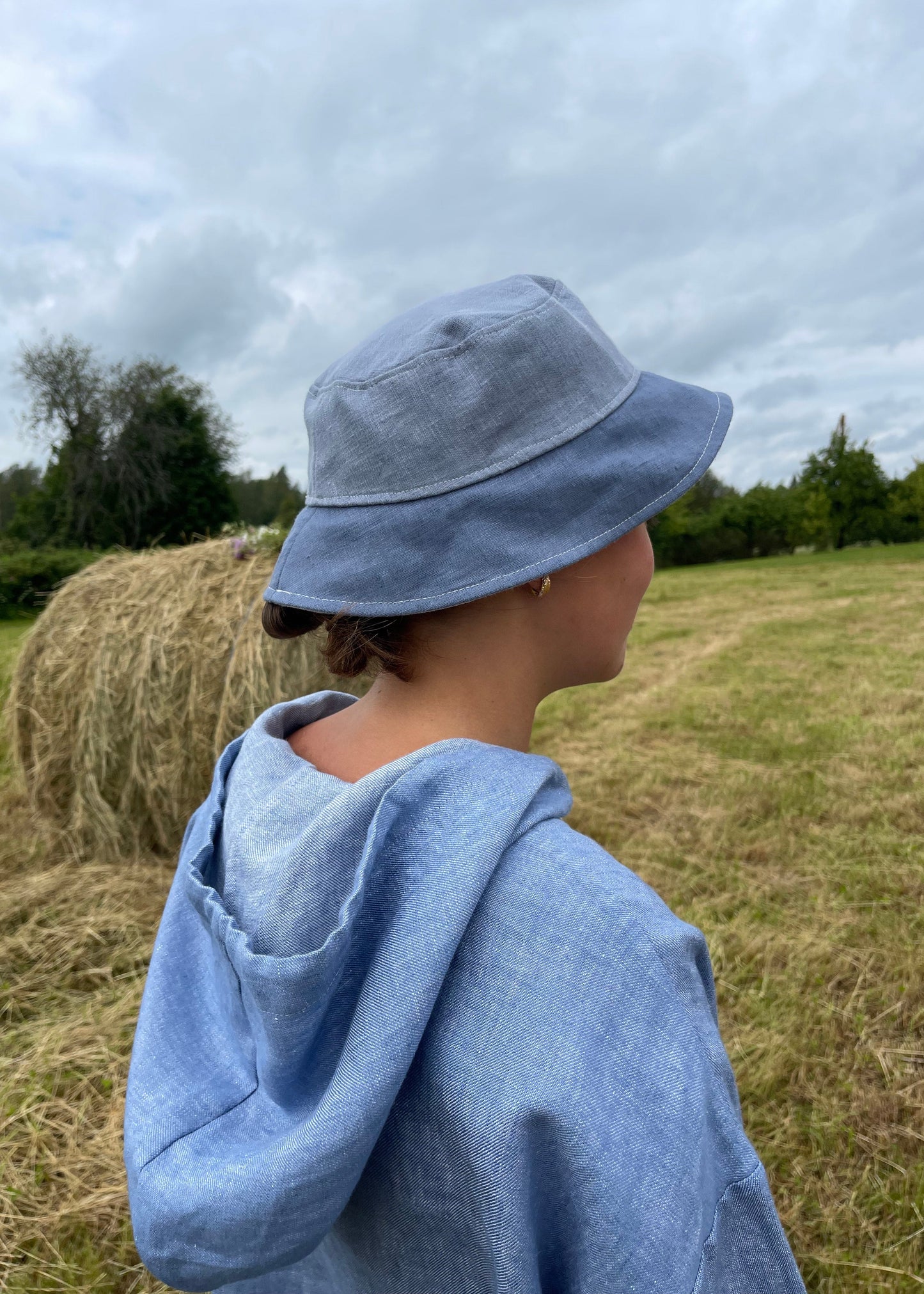 Person wearing a blue hoodie and hat standing in a field with hay bales.