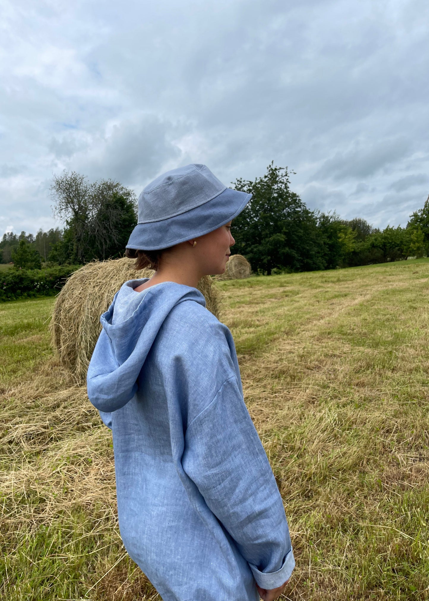 Person wearing a blue sweatshirt and cap standing in a grassy field with a cloudy sky.