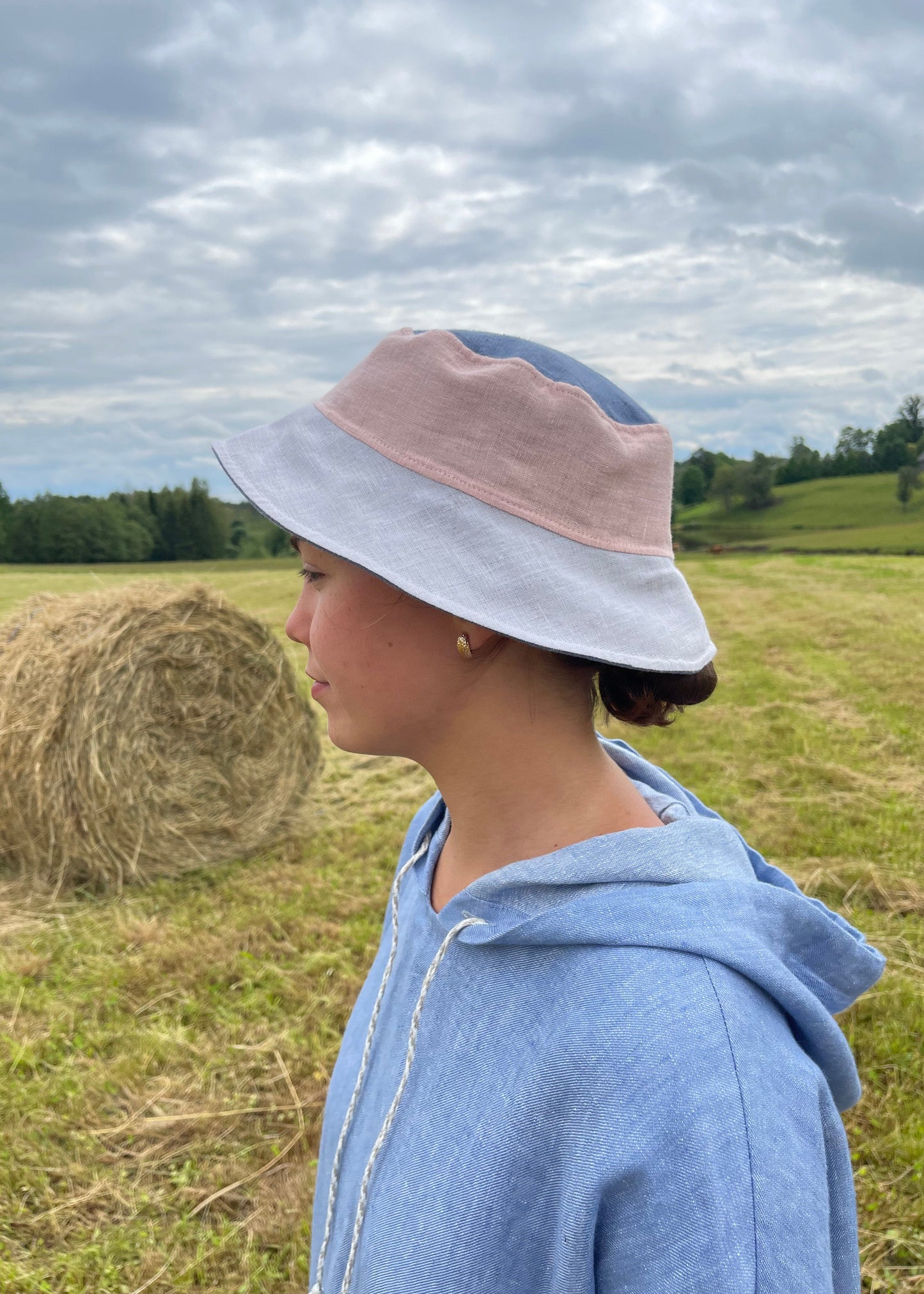 Person wearing a blue jacket and light gey hat in a field with a hay bale.